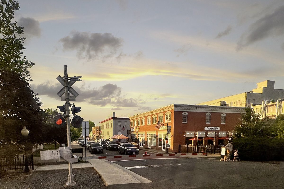 Downtown Manassas at sunset, with a railroad crossing signal in the foreground, parked cars and a corner brick restaurant with red umbrellas under a golden sky