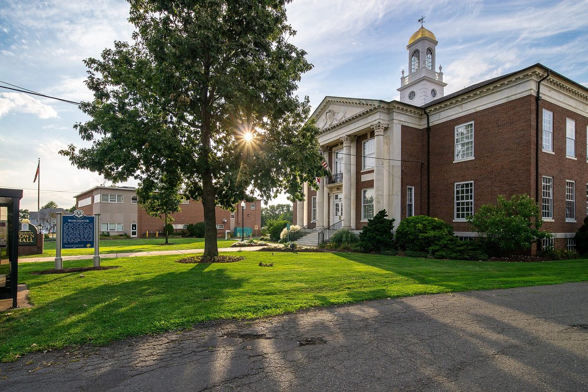 Manchester Town Hall at sunset: a red brick building with white columns and a gold-domed clock cupola, a large tree and a blue historical marker on the lawn