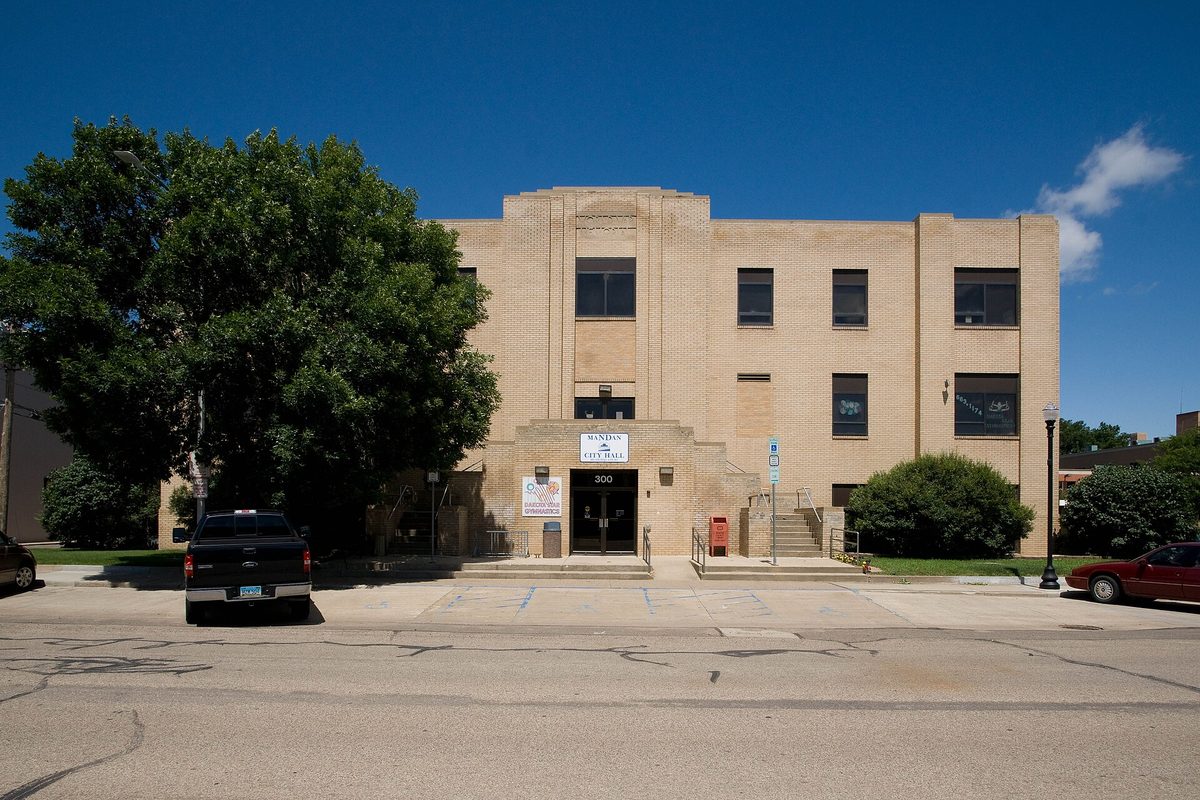 Mandan City Hall, a three-story tan brick building with a black pickup and a red car parked out front on a sunny day