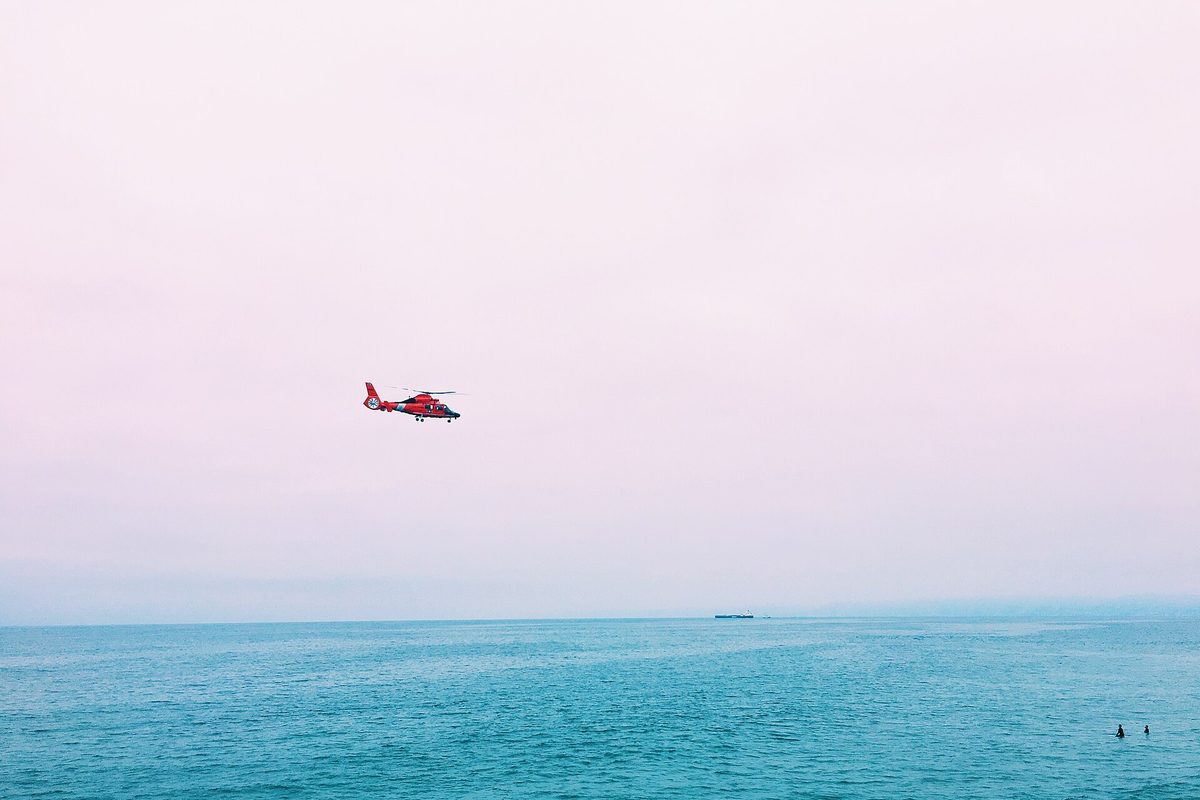 A red and white helicopter flying low over blue-green ocean water off Manhattan Beach under a hazy pink sky
