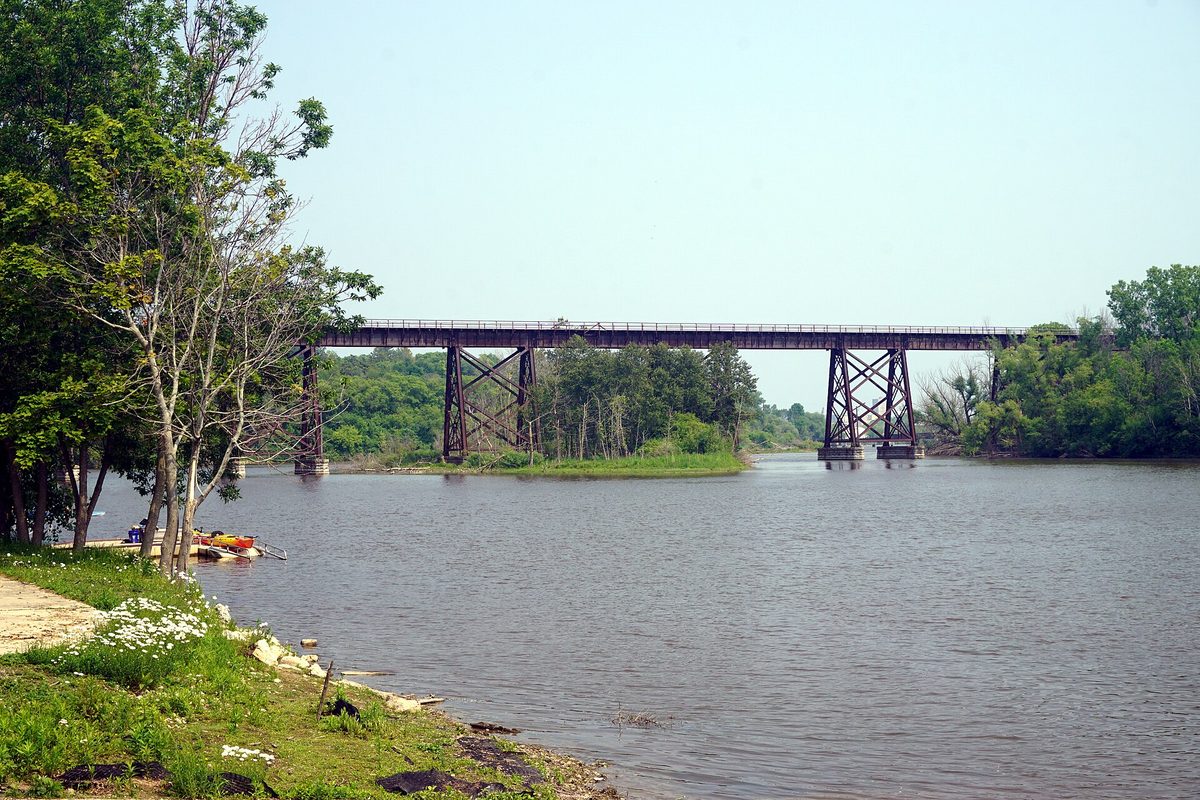 The Manitowoc River at Manitou Park, with a steel railroad trestle crossing the water and kayaks pulled up on the grassy bank