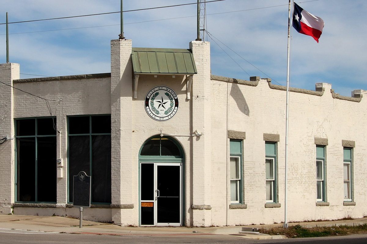 Manor's former city hall, a whitewashed brick building with a green metal awning over an arched glass door, the city seal on its front and a Texas flag flying beside it
