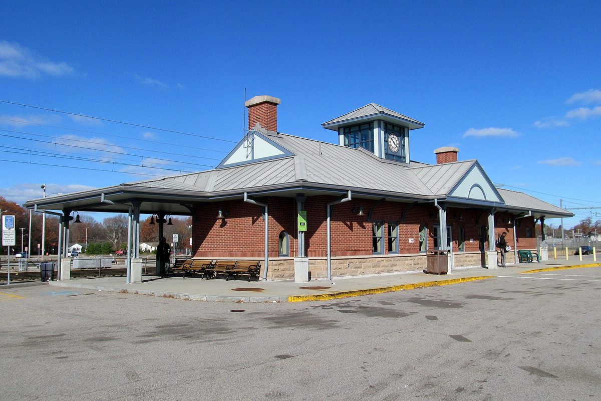 The red brick Mansfield commuter rail station with a gray metal roof, a clock cupola and benches under the canopy, on a clear blue day