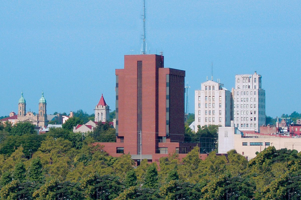 Mansfield's skyline above a band of green treetops: a tall brick tower with a radio mast in the center, two green church domes and a red spire at left, pale high-rises at right