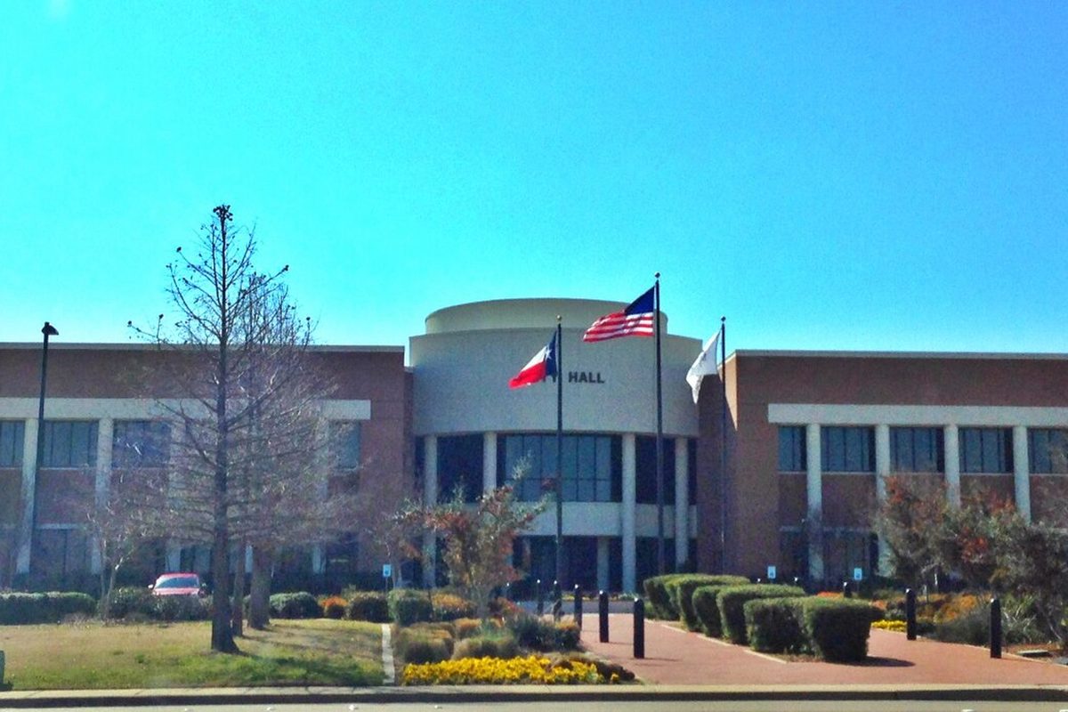Mansfield City Hall, a brick building with a rounded front entrance, with U.S., Texas and city flags flying over trimmed hedges