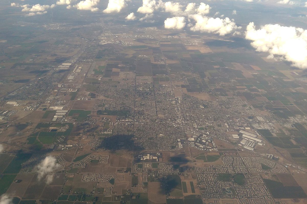 Aerial view from high above Manteca and Lathrop, a grid of streets and subdivisions surrounded by flat farmland under scattered white clouds