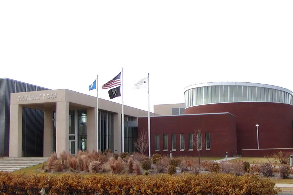 The Maple Grove Government Center, a tan stone entrance beside a round red-brick building with a glass band on top, three flags and autumn shrubs in front