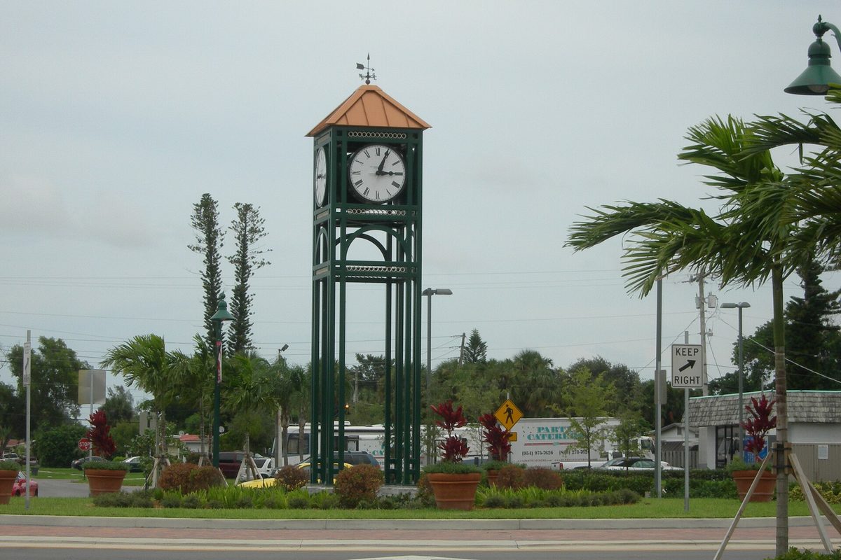 Margate's green clock tower with a copper-colored roof and weather vane, standing in a planted median with palms and flower pots