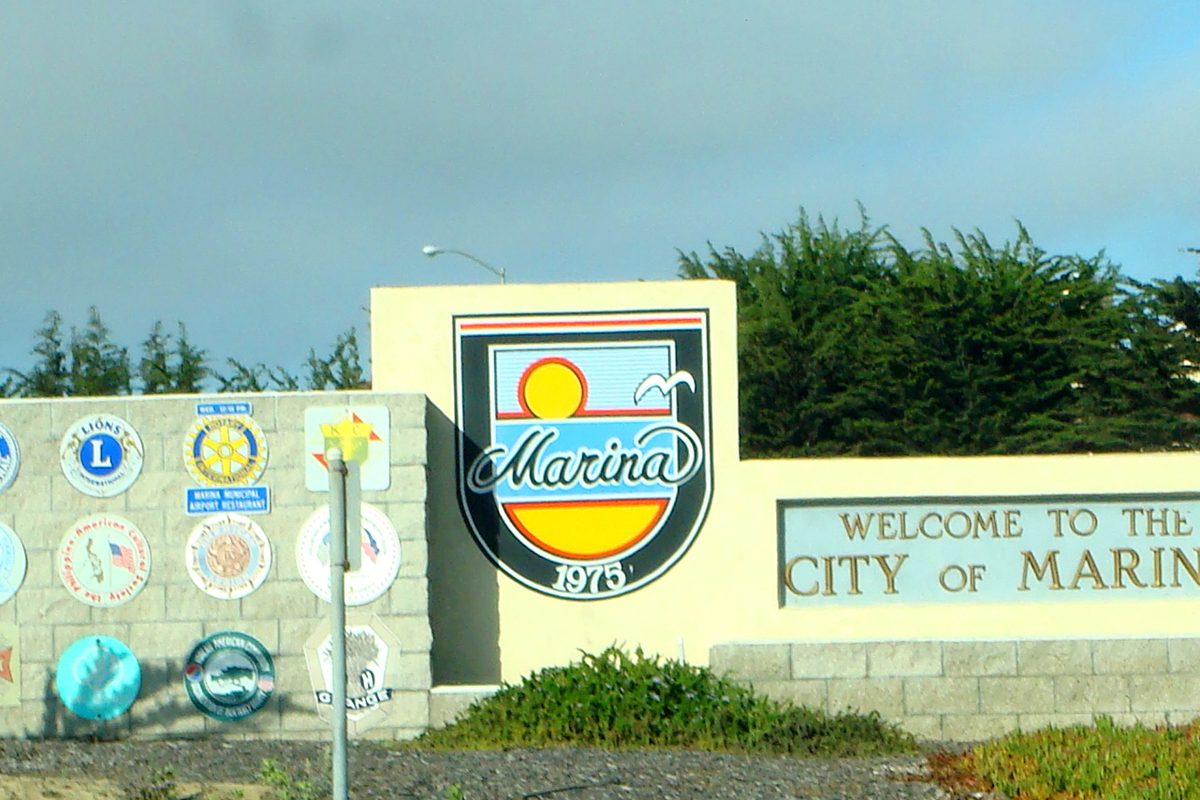 City of Marina welcome sign with the city crest dated 1975, beside a wall of service club emblems and cypress trees