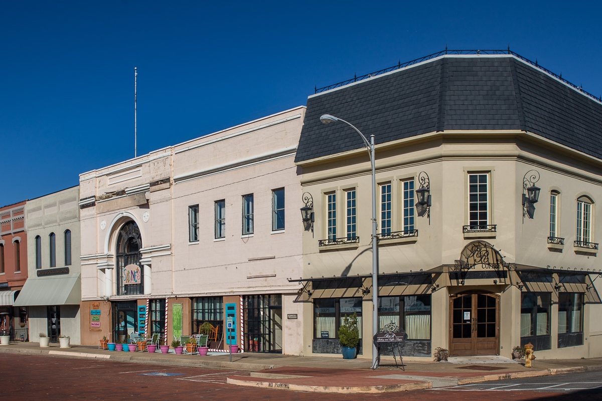 A large yellow-brick building with white columns, a central dome and a statue on its roof, with a white pickup parked on the red brick lot in front
