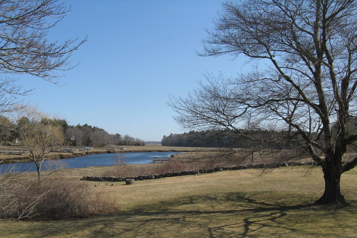 The North River curving through open marsh in Marshfield, with a dry lawn, a low stone wall and a large bare tree in the foreground under a clear sky