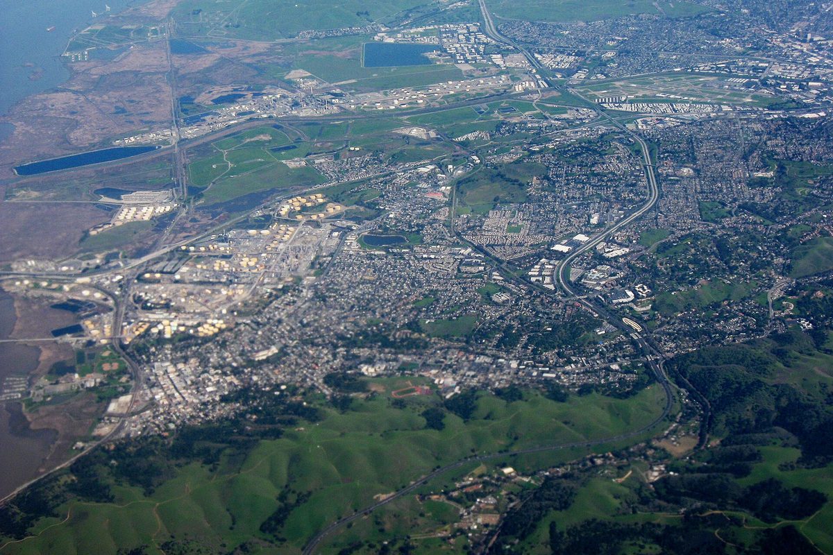Aerial view of Martinez facing east, with the Carquinez Strait and shoreline marsh on the left, green hills in front and Highway 4 curving up the right side