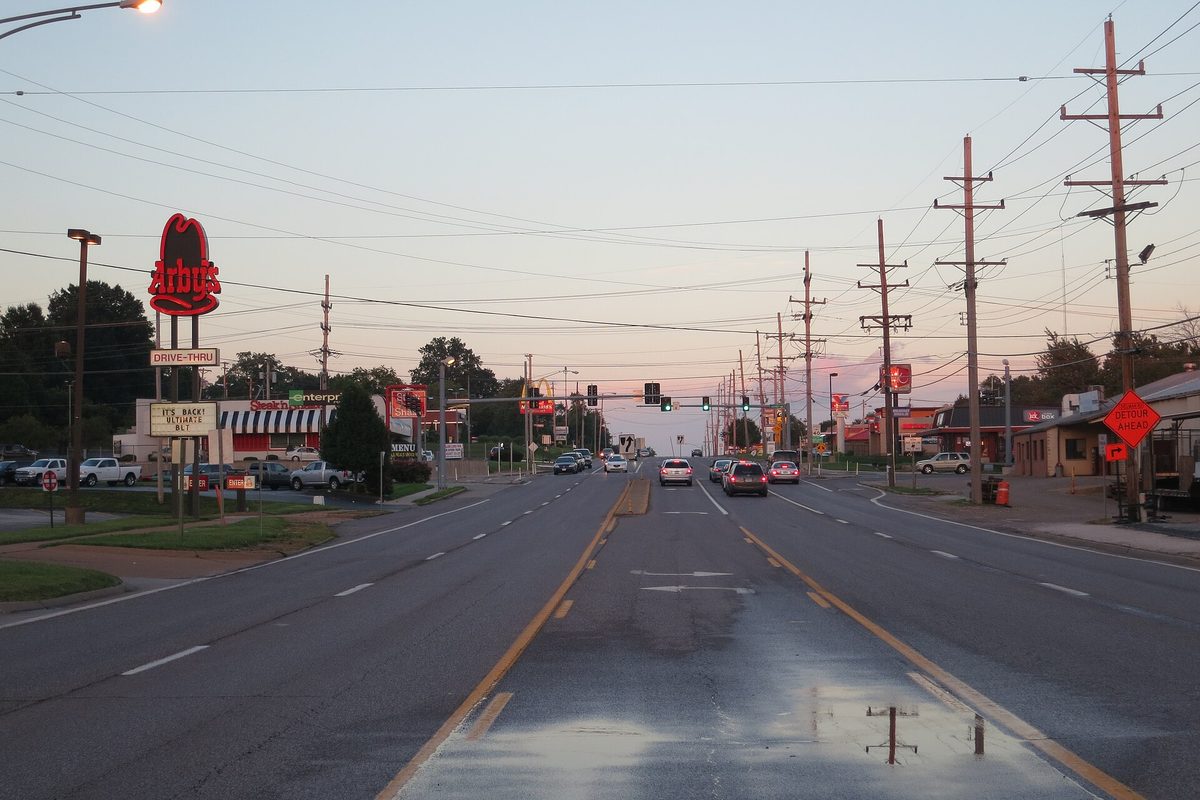 Page Avenue in Maryland Heights at dusk, a wet four-lane road lined with traffic lights, utility poles and a lit fast-food sign