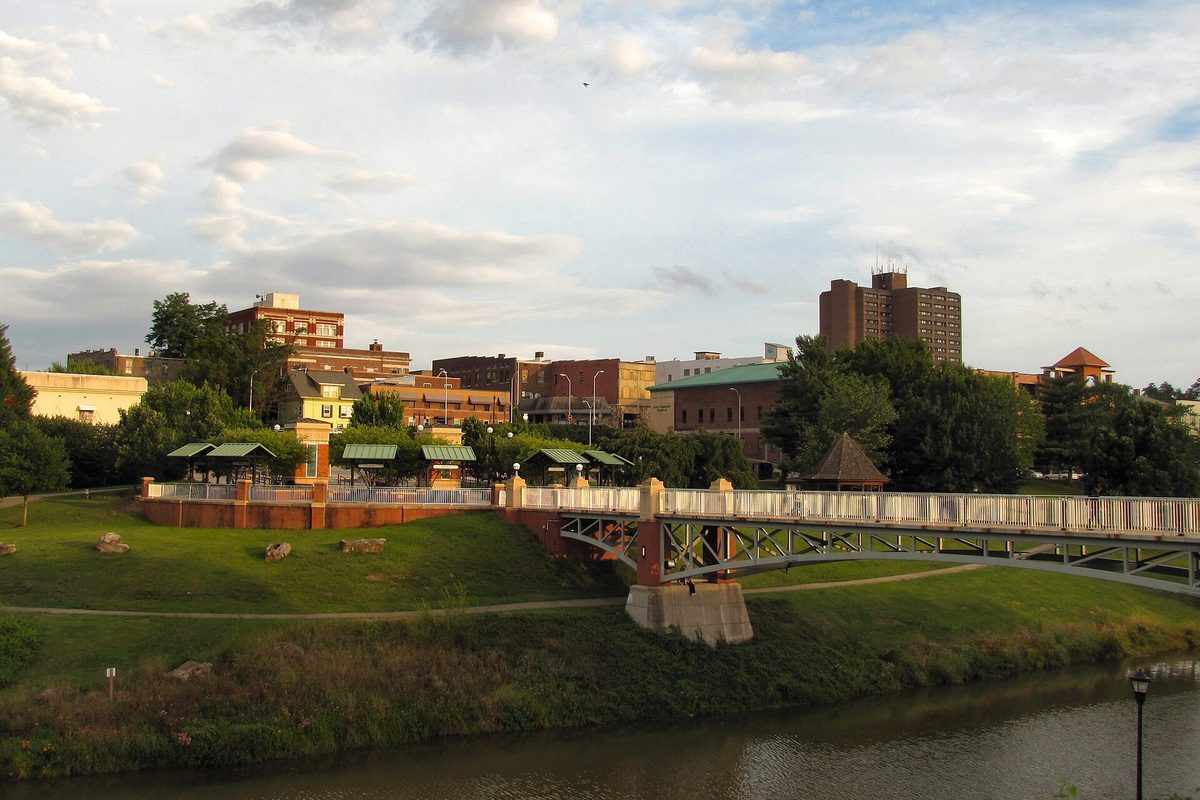 Maryville seen across Greenbelt Lake, with a footbridge, park pavilions on a grassy bank and brick buildings behind under a cloudy evening sky