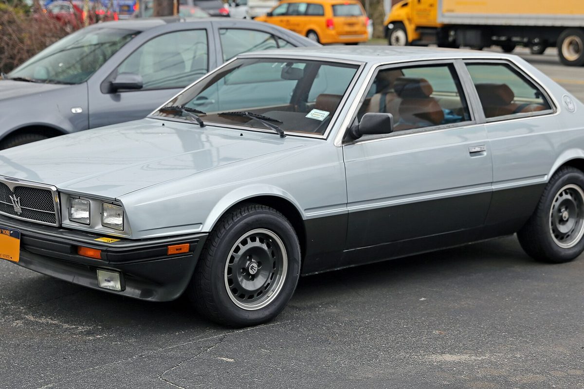 A silver Maserati Biturbo two-door coupe with a dark gray lower body and slotted wheels, parked in a lot beside other cars