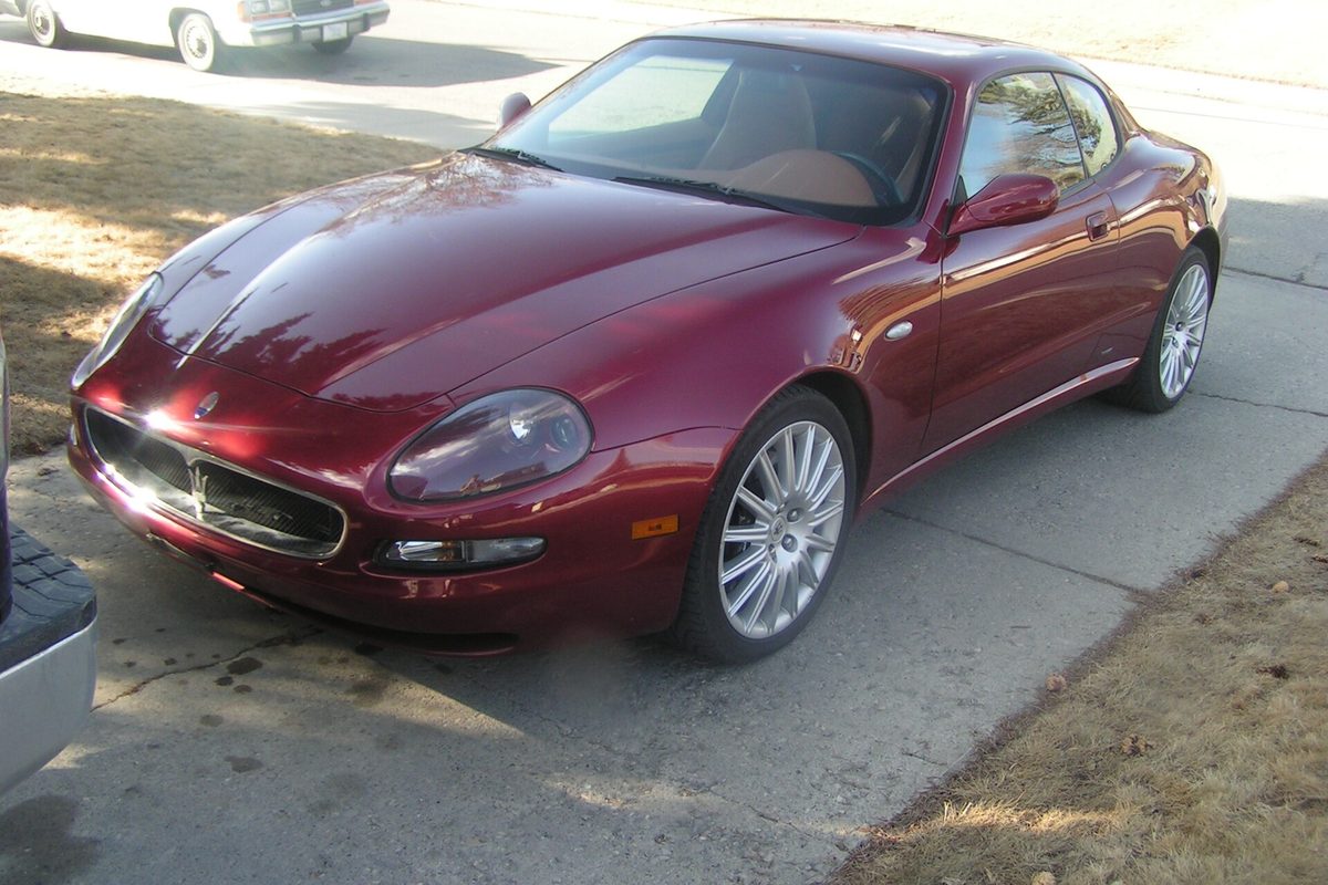 A dark red Maserati Coupe with silver multi-spoke wheels, parked on a concrete driveway in low winter sun