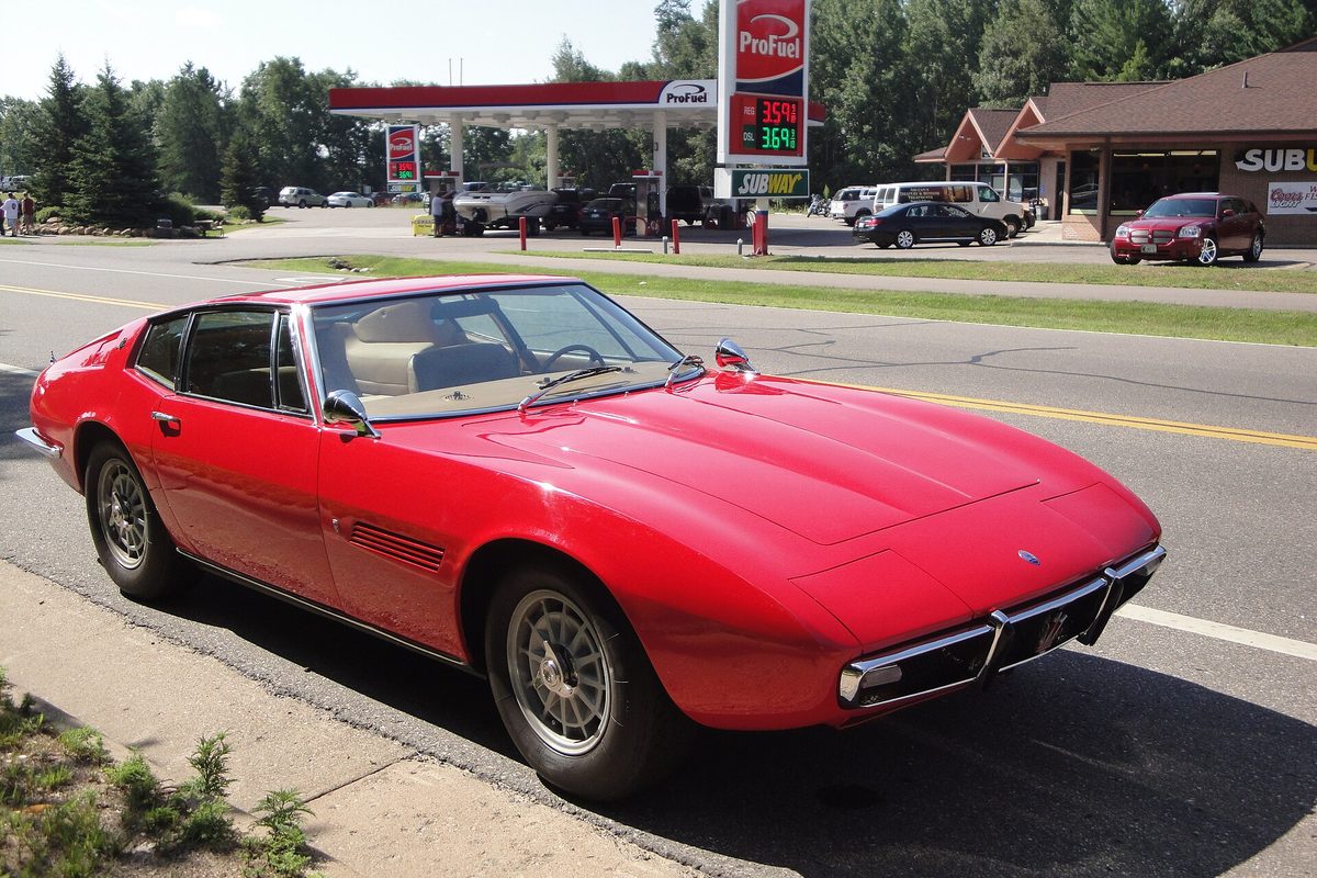 A red 1967 Maserati Ghibli fastback coupe parked at a roadside curb with a gas station behind it