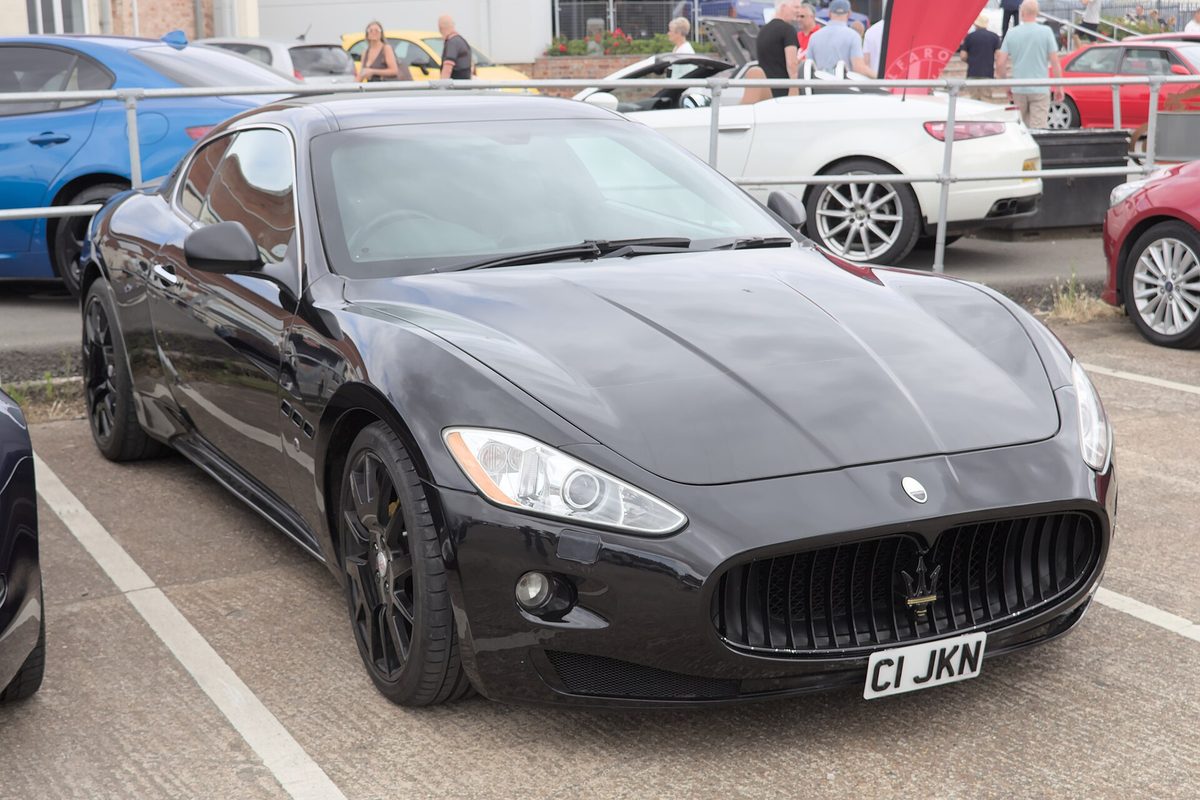 A black first-generation Maserati GranTurismo coupe with black wheels parked at an outdoor car show