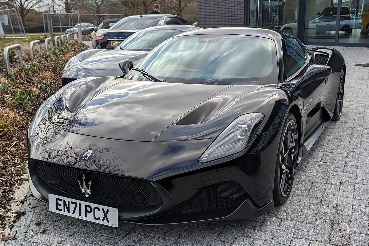 A black Maserati MC20 coupe parked on brick paving outside an office building, with its hood vents and grille in view