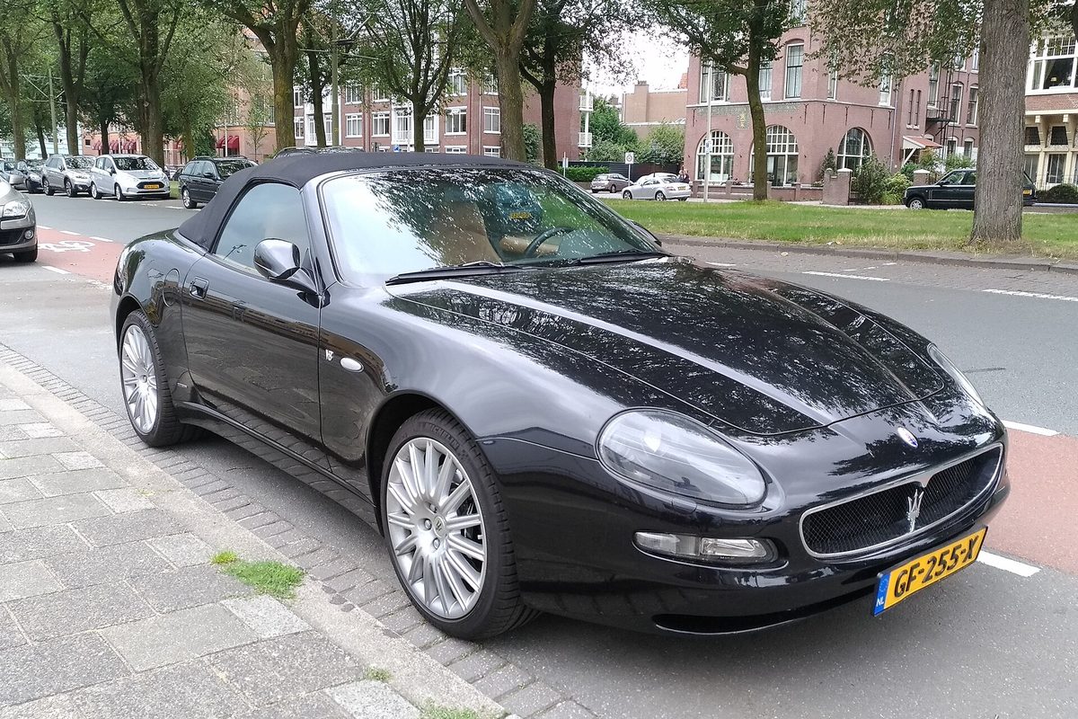 A black Maserati Spyder convertible with its soft top up, parked at a tree-lined city curb