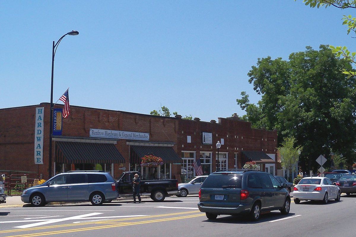Renfrow Hardware's long brick storefront in downtown Matthews, with a vertical Hardware sign, American flags and cars passing on the street