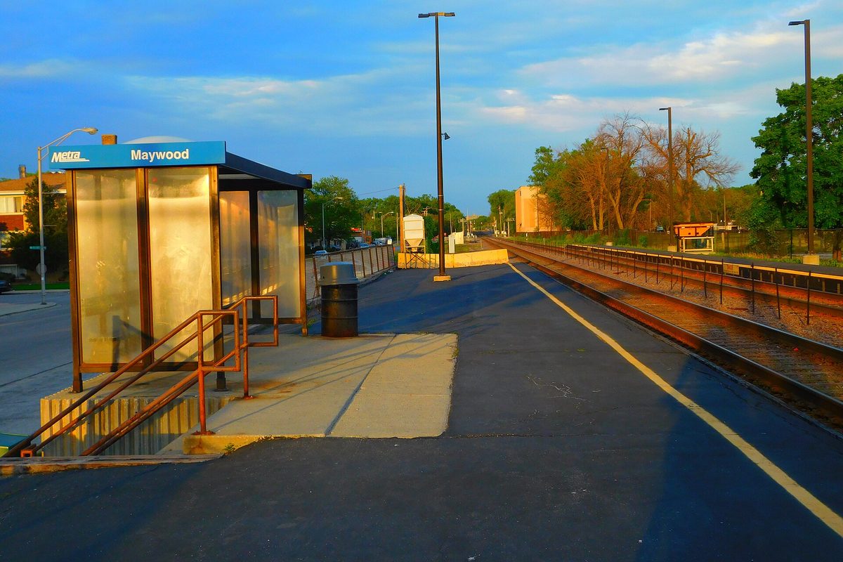 The Maywood Metra station platform at sunset, with a glass shelter under a blue Metra Maywood sign and the tracks running into the distance