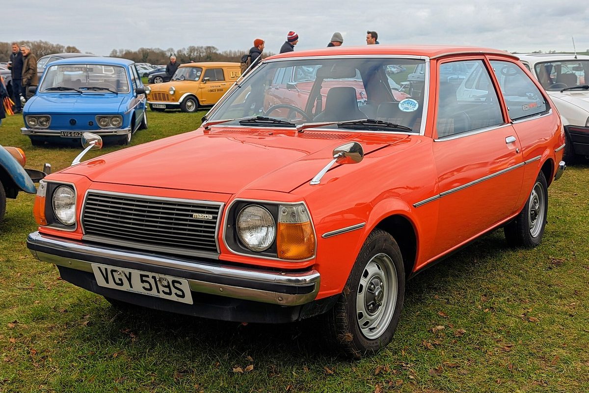 An orange 1978 Mazda 323 three-door hatchback with chrome bumpers and round headlights, parked on grass at a car show