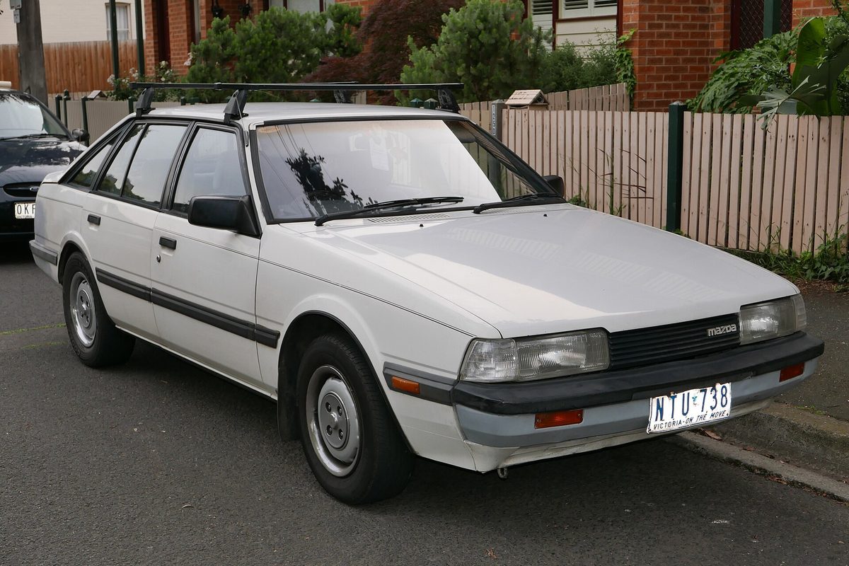 A white early-1980s Mazda 626 hatchback with a roof rack, parked at a curb beside a wooden fence