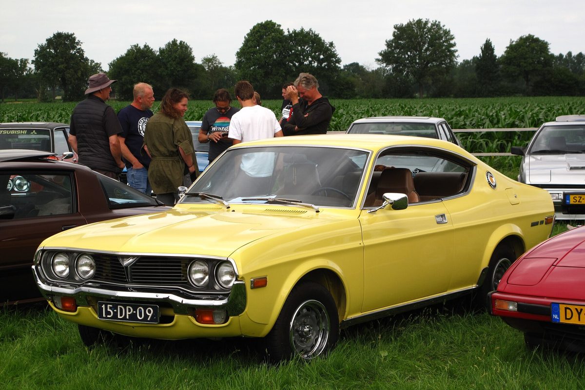 A yellow 1970s Mazda 929 coupe with four round headlights, parked on grass at a classic car show
