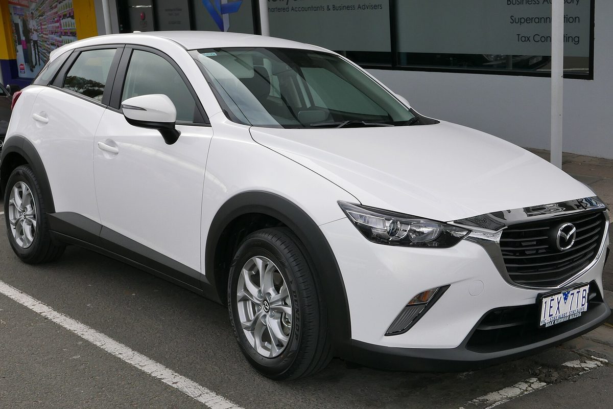 A white Mazda CX-3 small crossover with black wheel-arch trim and silver alloy wheels, parked at a curb outside a shopfront
