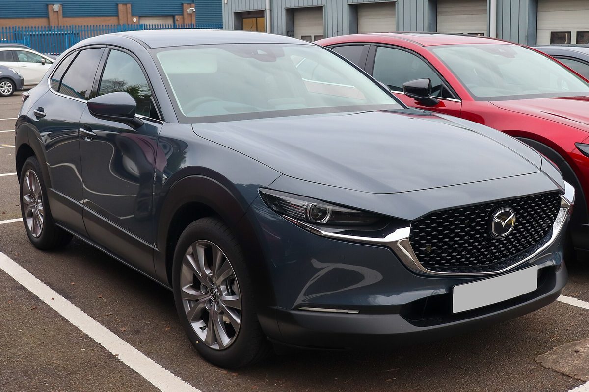 A dark blue-gray Mazda CX-30 crossover seen from the front corner, parked in a lot beside a red car