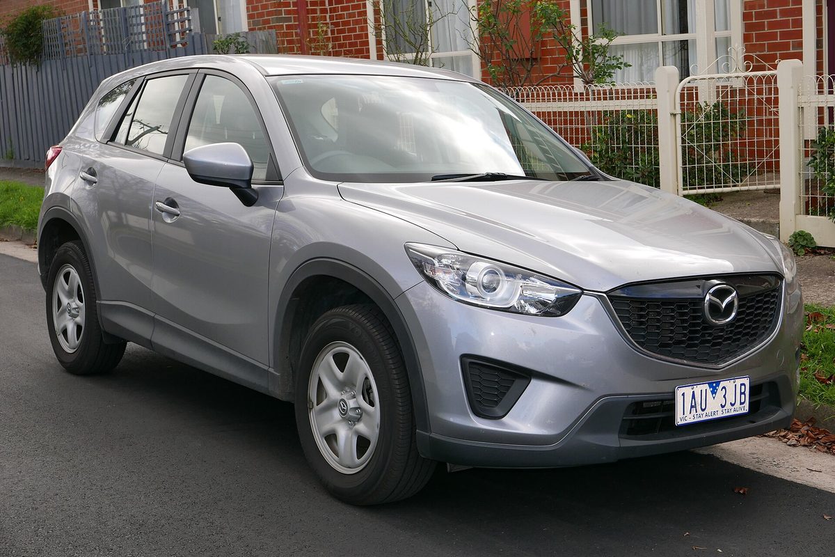 A silver first-generation Mazda CX-5 parked at a curb in front of a brick house and a white picket fence