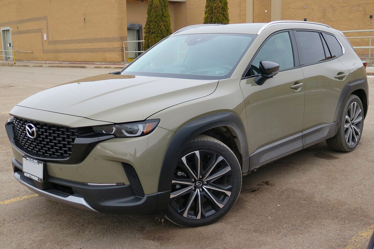 A 2023 Mazda CX-50 GT compact SUV in Zircon Sand, a muted green-beige, with black wheel arches and multi-spoke alloy wheels, parked in an empty lot