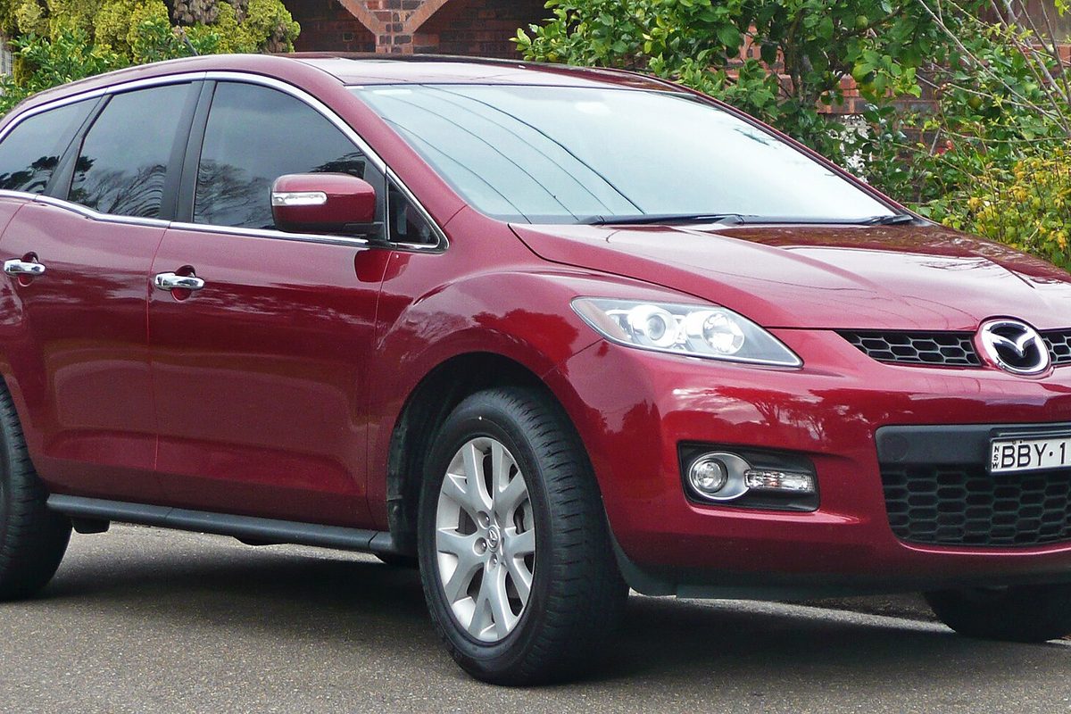 A red Mazda CX-7 crossover parked on a street, seen from the front right, with green shrubs and a brick wall behind it