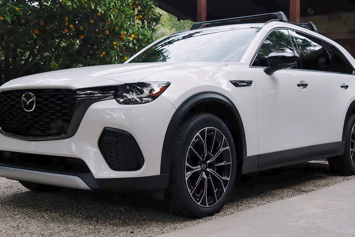 A white Mazda CX-70 SUV with black roof rails and two-tone alloy wheels, parked on a gravel drive beside an orange tree