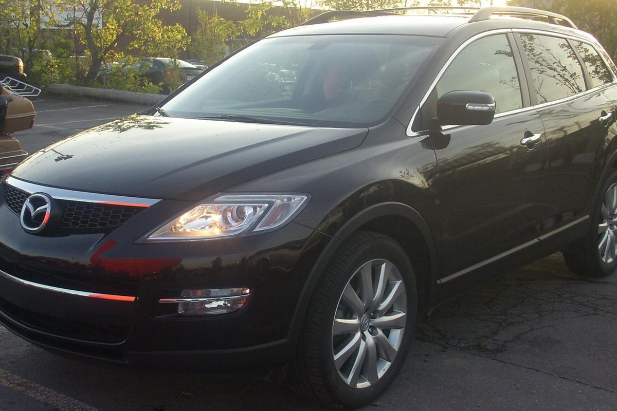 A black first-generation Mazda CX-9 with its headlights on, parked in a lot in low evening sun