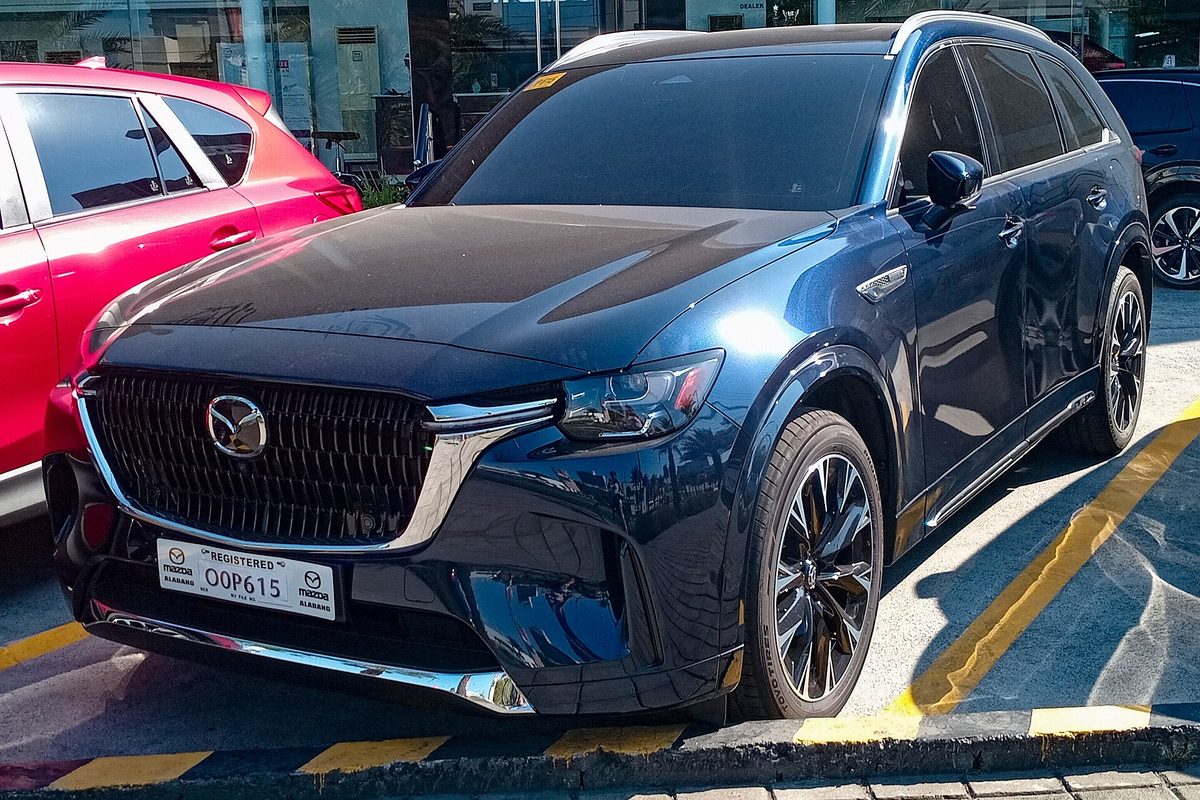A dark blue Mazda CX-90 SUV parked in a lot with yellow lines, seen from the front left, next to a red car