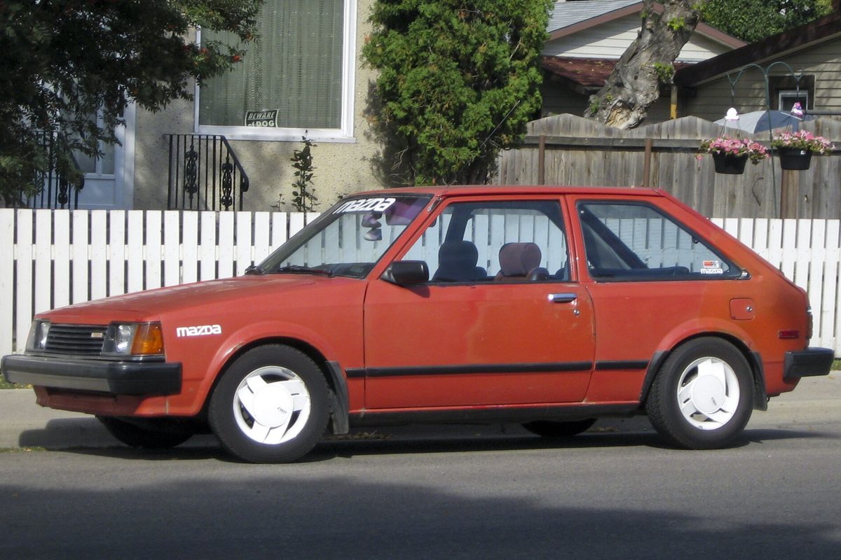 A faded red Mazda GLC three-door hatchback with white wheel covers, parked at a curb in front of a white picket fence