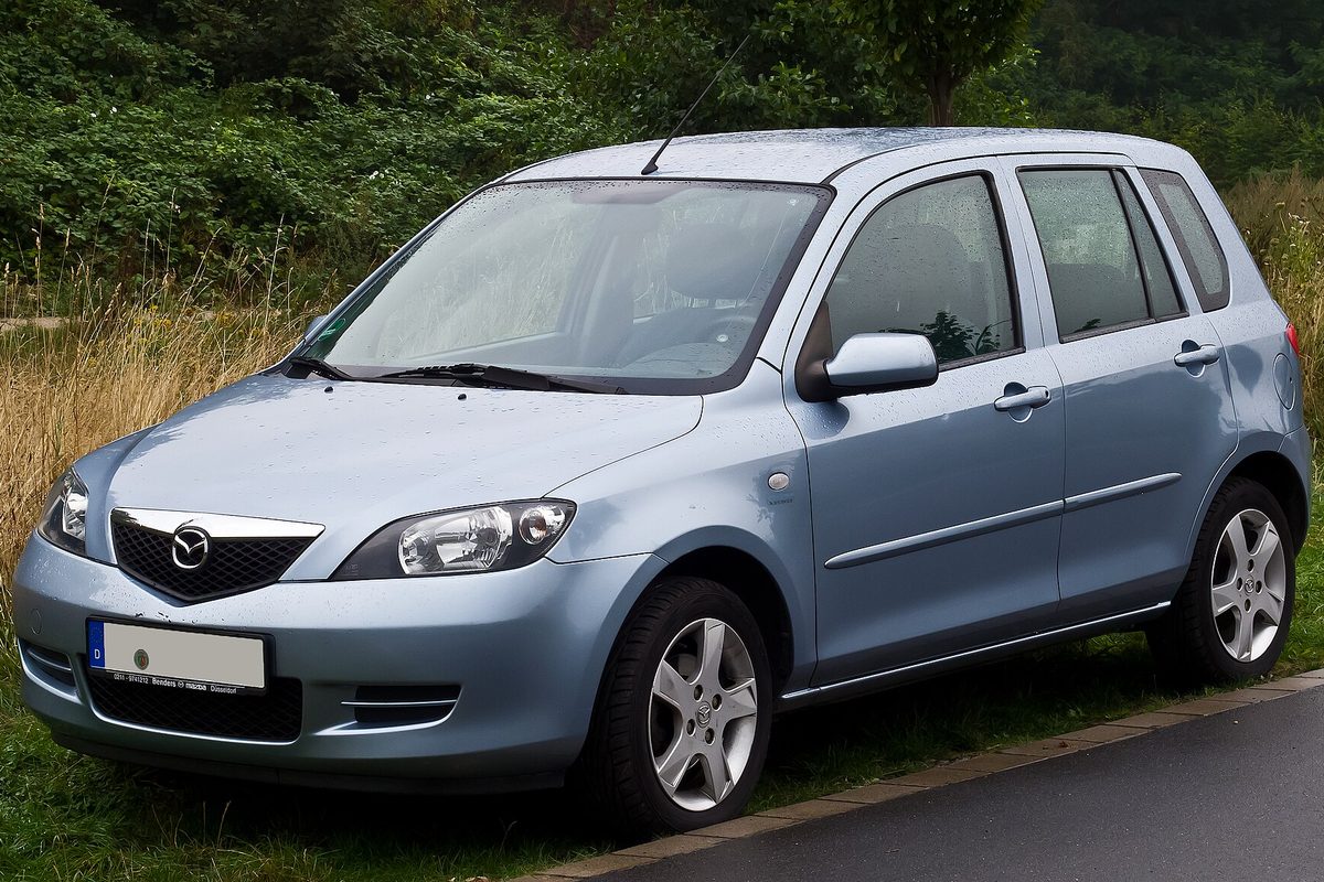 A light blue Mazda2 five-door hatchback parked on wet grass at the edge of a road, with tall grass and trees behind it