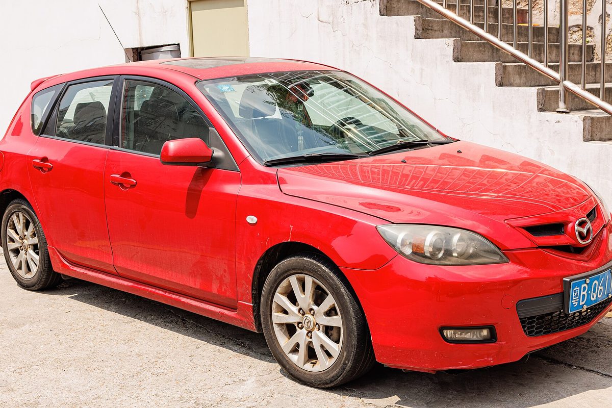 A red first-generation Mazda3 hatchback parked on a concrete lot below an outdoor stairway