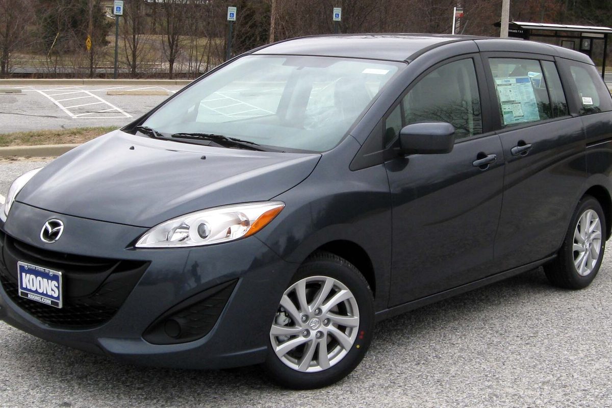 A dark gray Mazda5 compact van with a window sticker on its rear side glass, parked in a paved lot