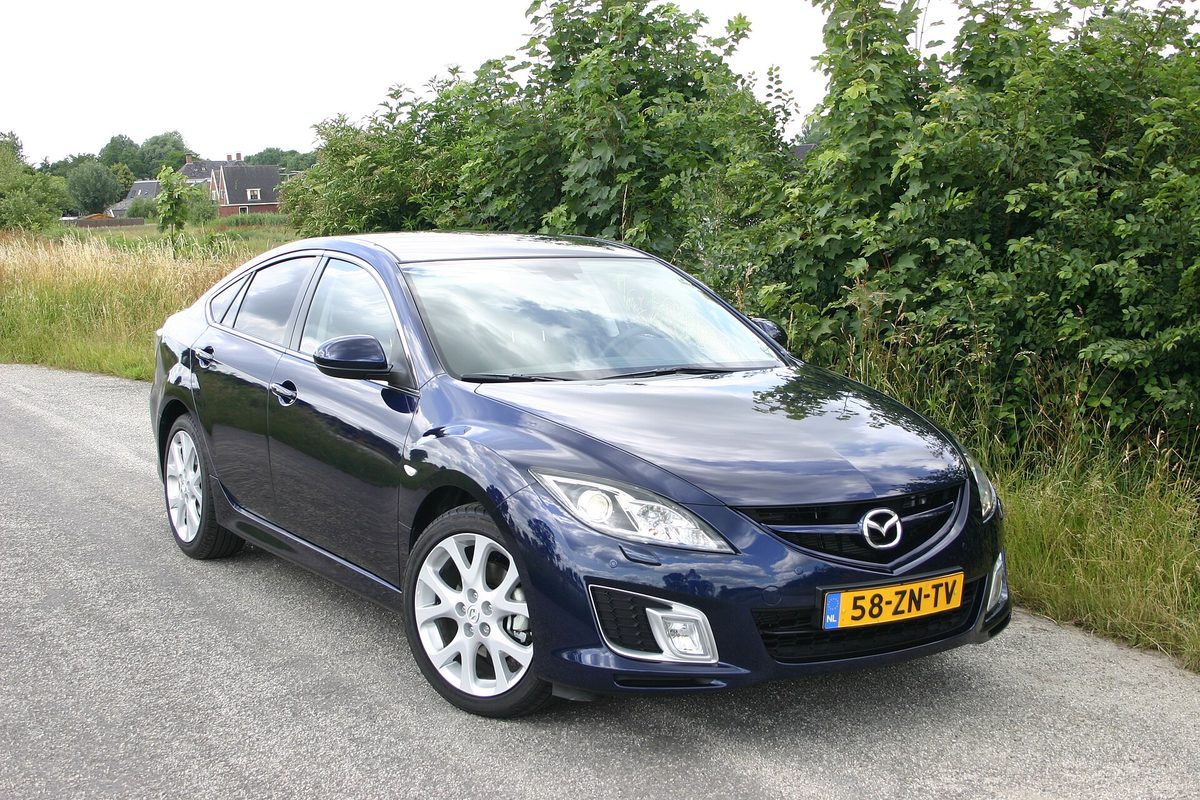 A dark blue Mazda6 hatchback parked on a narrow country road lined with tall green bushes