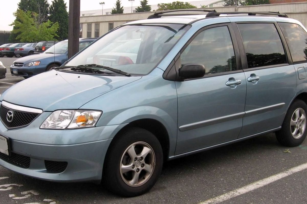 A light blue Mazda MPV minivan with roof rails, parked in a store lot with other cars behind it