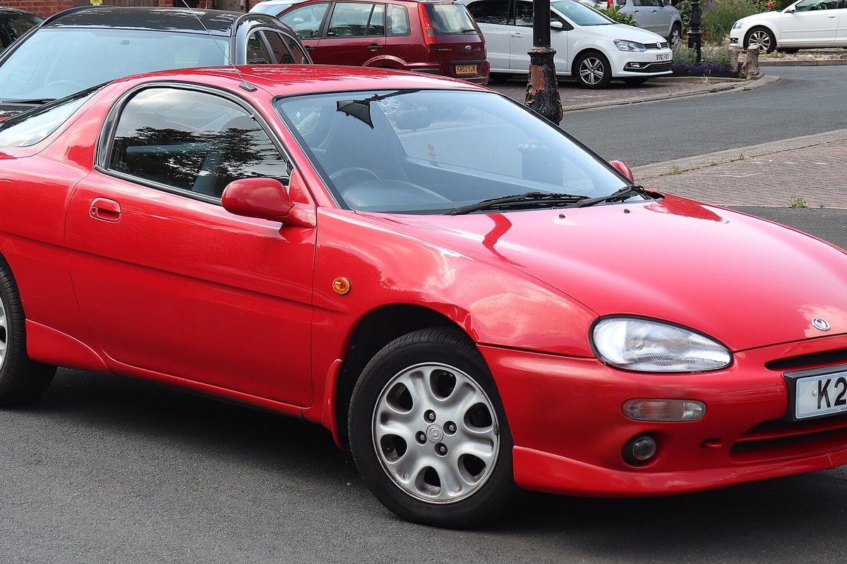 A bright red 1992 Mazda MX-3 hatchback coupe with silver alloy wheels, parked on a street with houses behind it