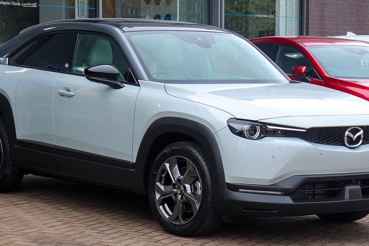 A pale grey Mazda MX-30 crossover with a black roof and black wheel arches, parked on brick paving outside a dealership