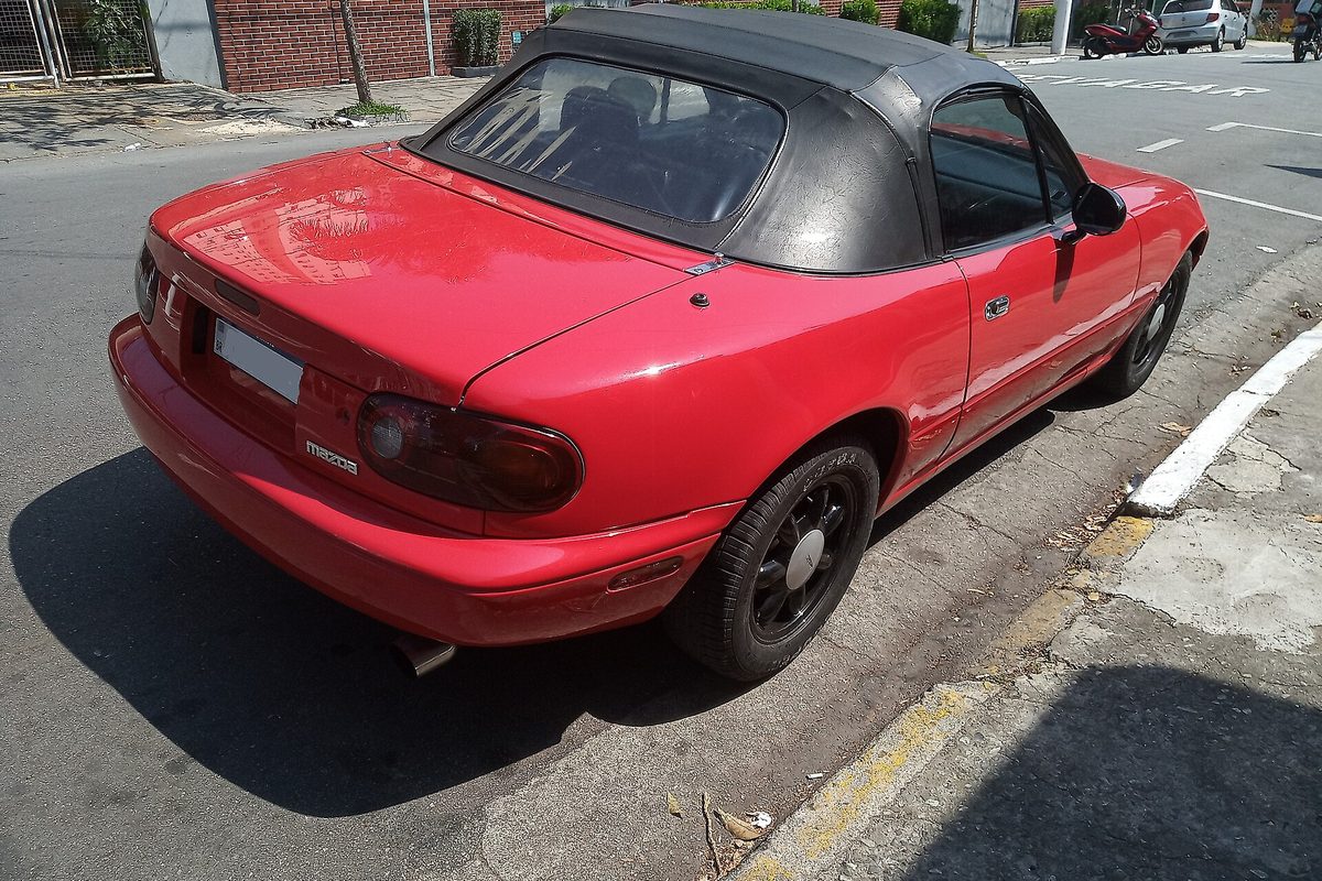 A red Mazda MX-5 Miata convertible with its black soft top up, seen from the rear corner, parked at a curb on dark wheels