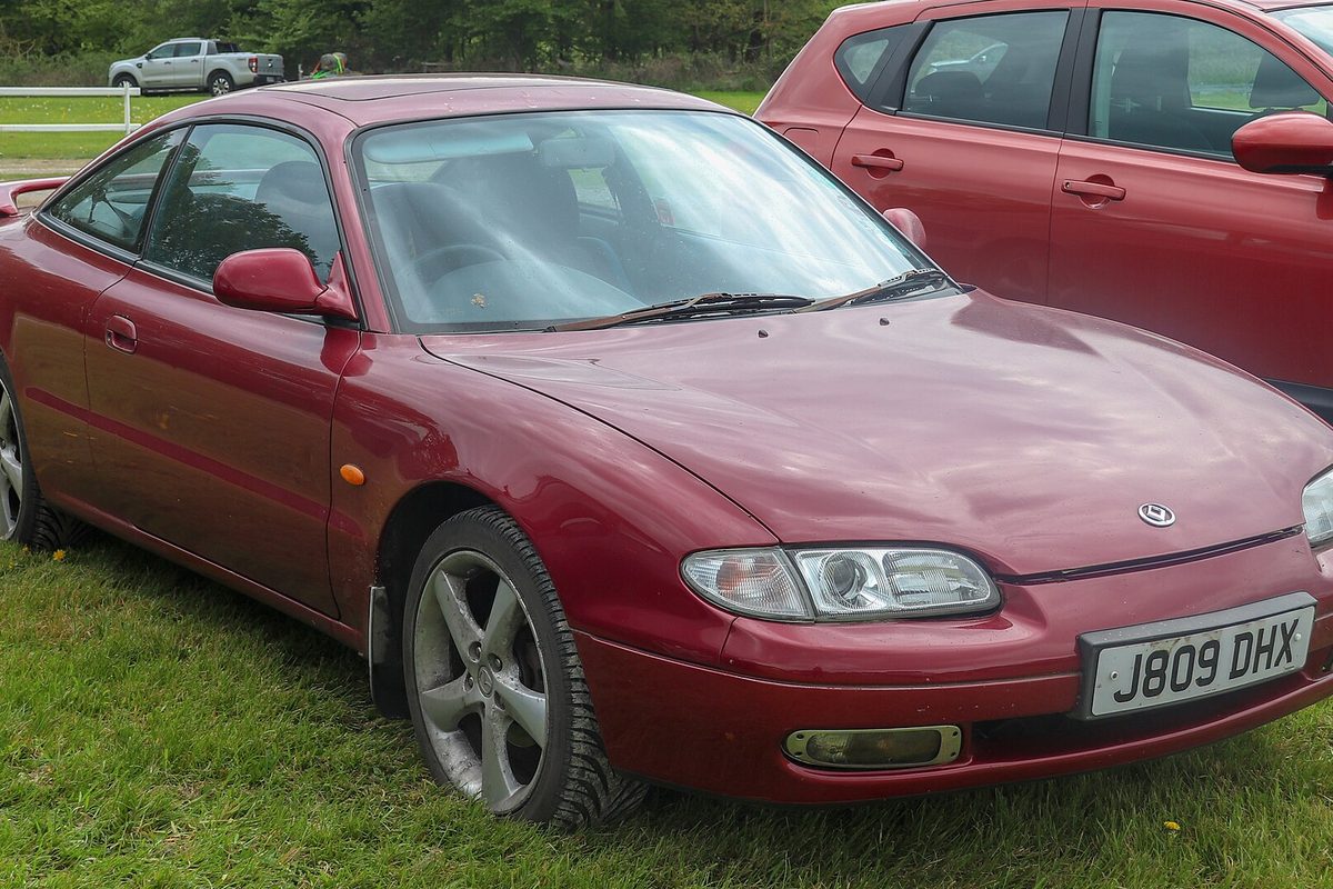 A dark red 1992 Mazda MX-6 V6 two-door coupe with silver five-spoke alloy wheels, parked on grass in front of a red hatchback