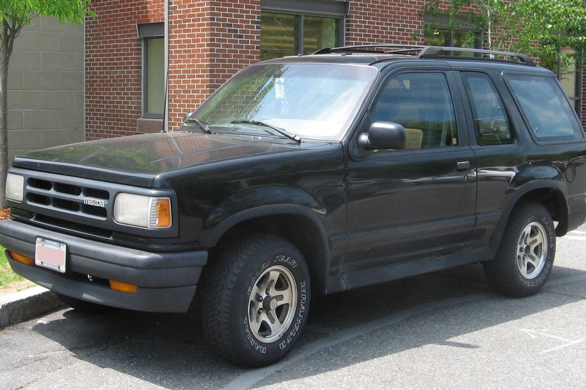 A black two-door Mazda Navajo SUV with a roof rack and silver alloy wheels, parked at a curb beside a brick building