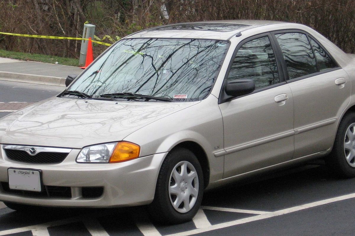A champagne Mazda Protege sedan parked in a lot, with yellow tape and an orange cone behind it