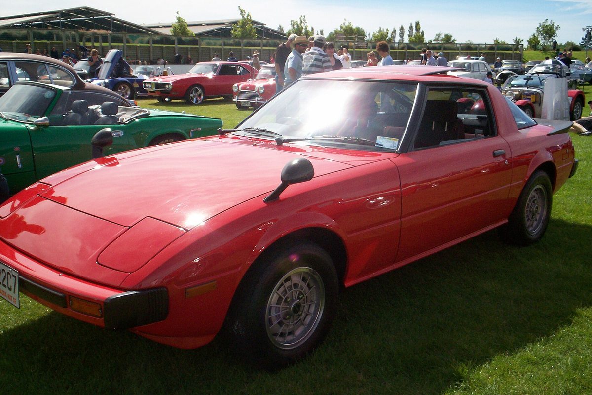 A red first-generation Mazda RX-7 coupe with mirrors on the hood, parked on grass at a car show among other classics